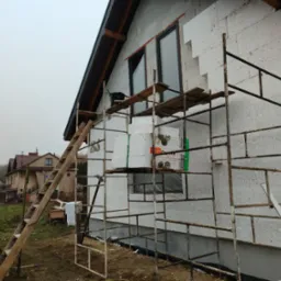 Budowa domu: ściany z białych bloczków, rusztowanie, okna z czarną ramą, styropian. Widok na dom w tle, pochmurne niebo.