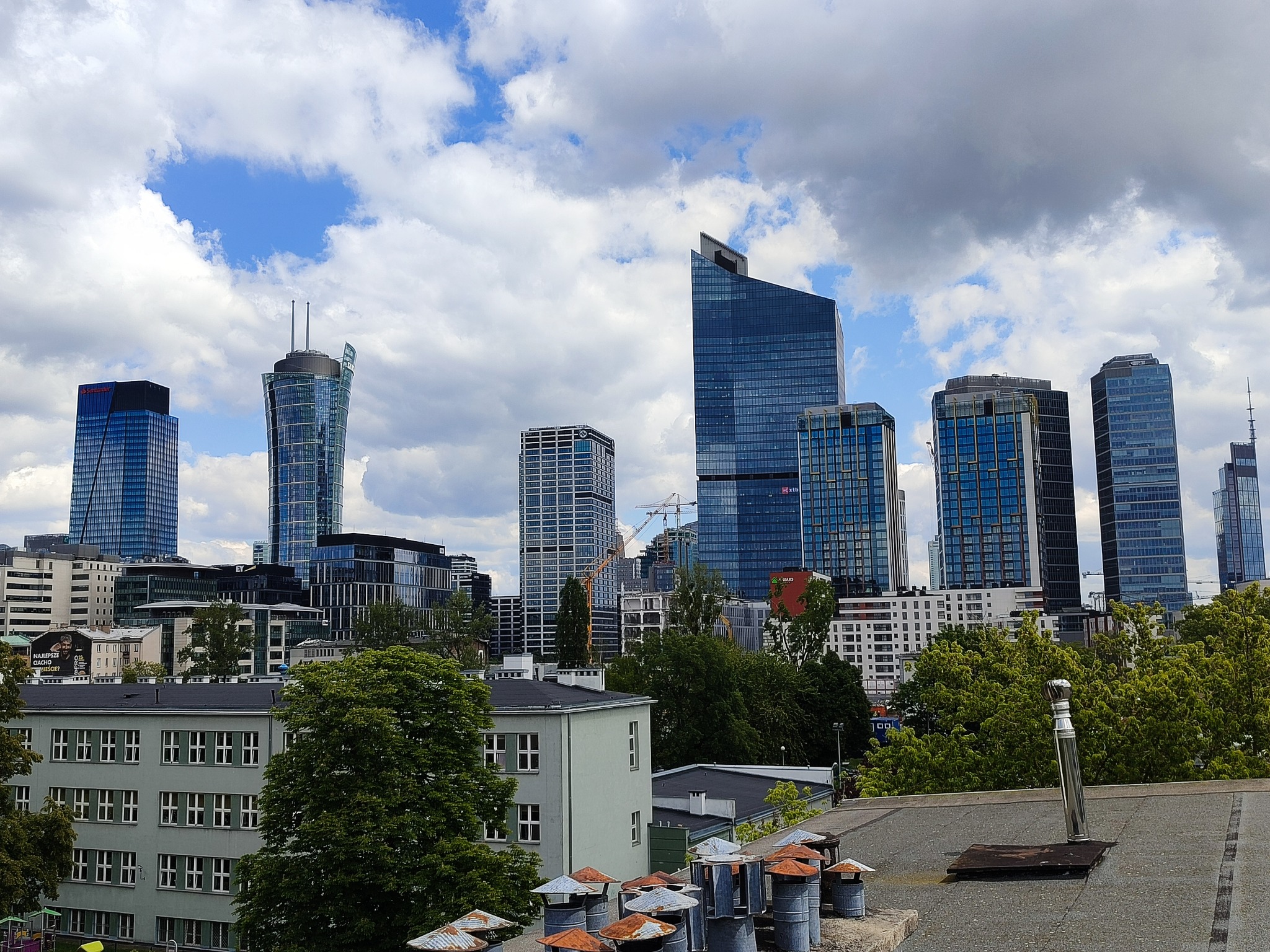 Panorama miasta z wysokimi budynkami, widok z dachu budynku z kominami, w tle błękitne niebo z chmurami.