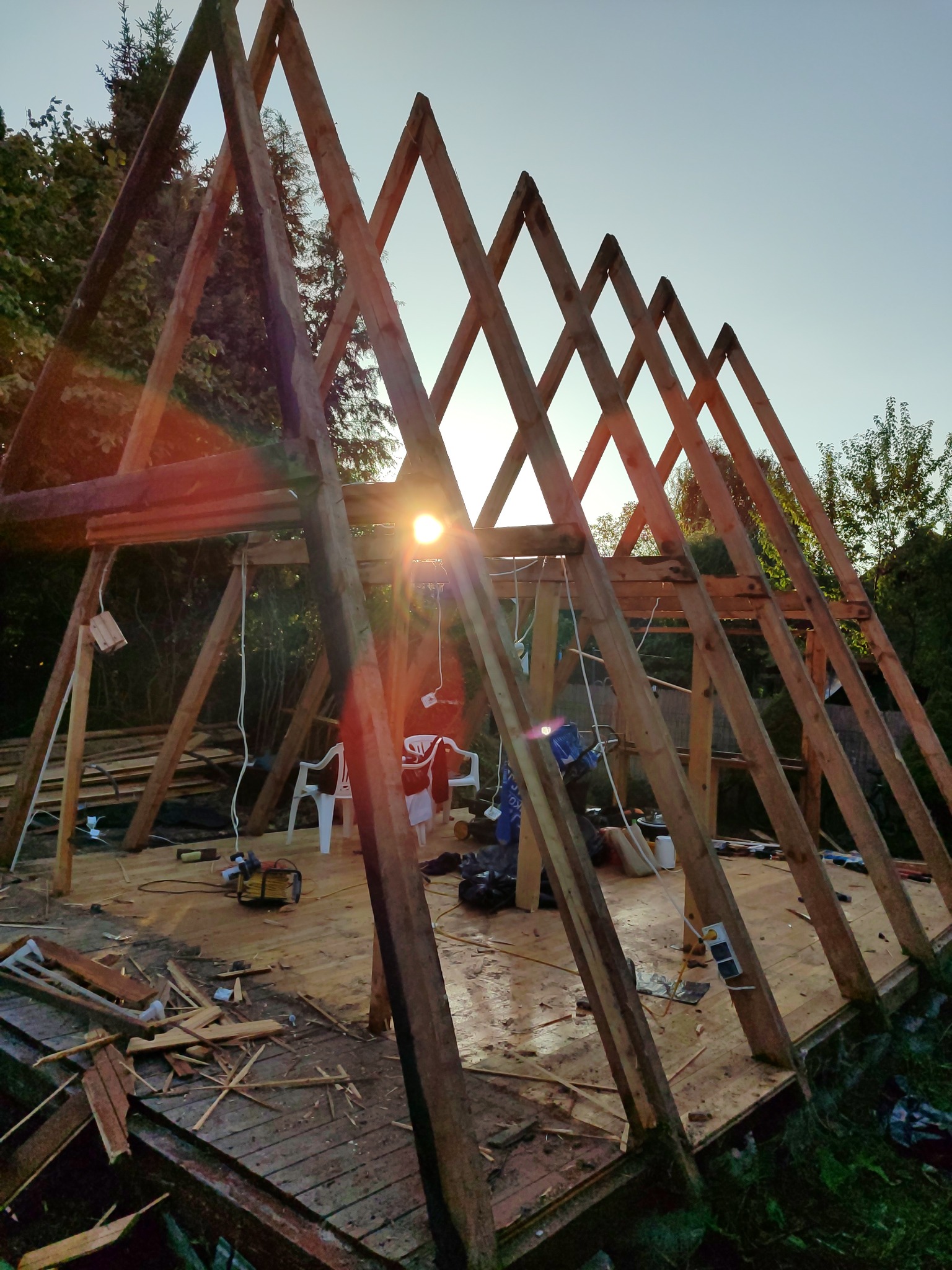 Drewniana konstrukcja w kształcie litery A w trakcie budowy, widoczne narzędzia i elementy konstrukcyjne. Słońce prześwieca przez szkielet, tworząc ciepłe światło.