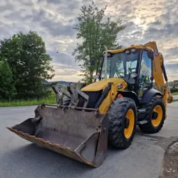 Żółta koparko-ładowarka JCB 4CX na asfaltowej drodze, z widocznym łyżką i ramieniem kopiącym, na tle zieleni i pochmurnego nieba. Maszyna gotowa do pracy.