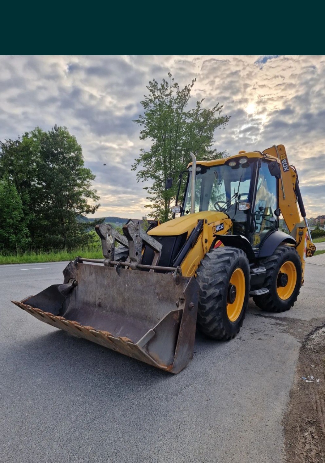 Żółta koparko-ładowarka JCB 4CX na asfaltowej drodze, z widocznym łyżką i ramieniem kopiącym, na tle zieleni i pochmurnego nieba. Maszyna gotowa do pracy.