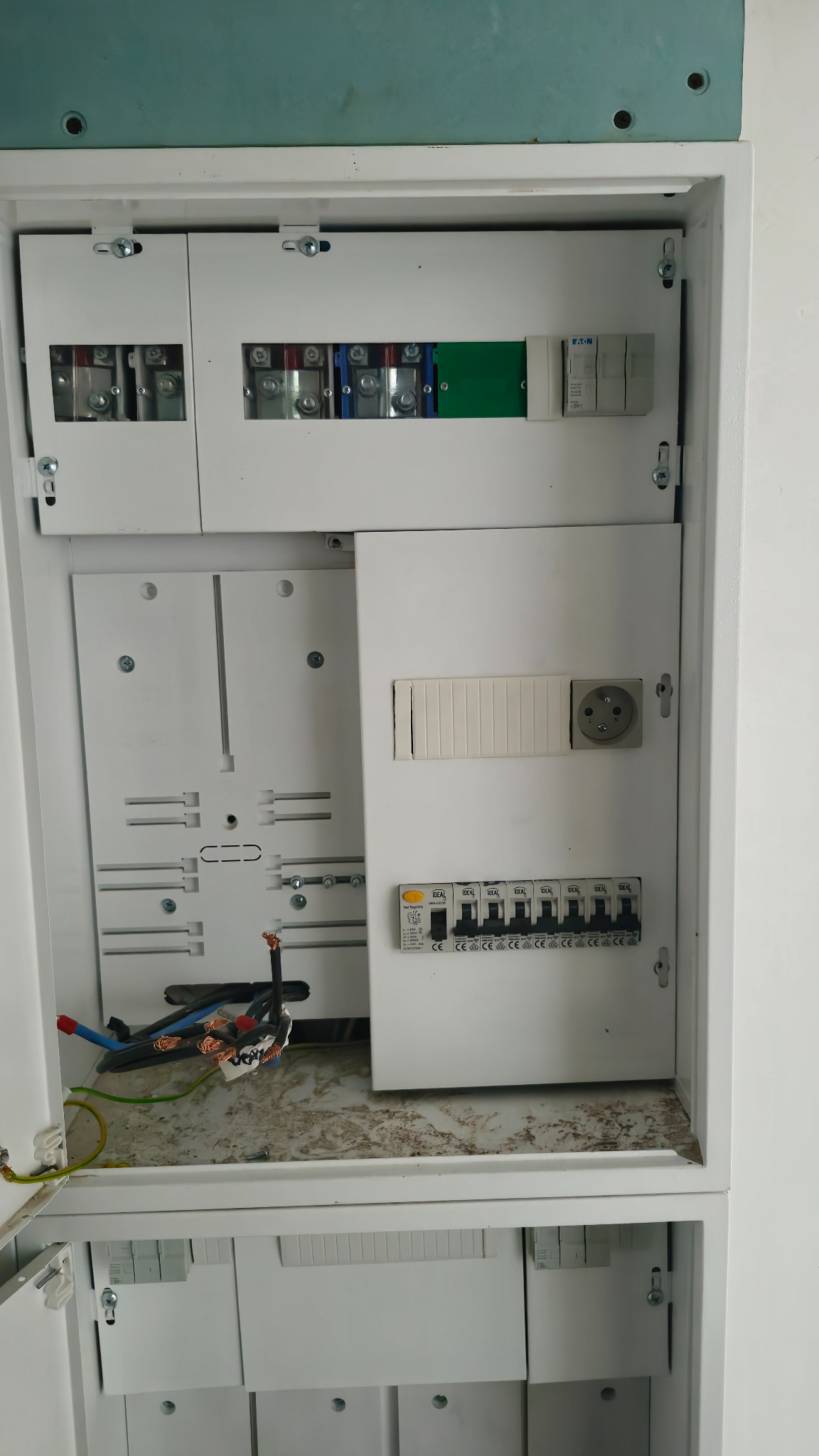 Otwarta, biała skrzynka rozdzielcza z widocznymi bezpiecznikami, gniazdem i przewodami. Widoczne elementy instalacji elektrycznej wewnątrz obudowy.