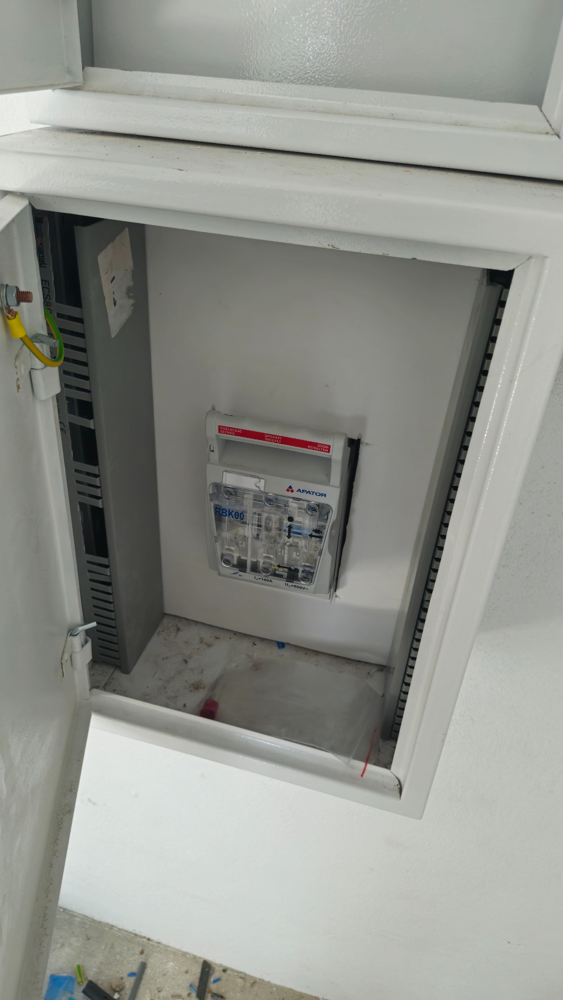 Otwarta, biała skrzynka elektryczna z zamontowanym bezpiecznikiem RBK00 firmy Apator. Widoczne przewody i szyny DIN. Ujęcie z góry, wnętrze skrzynki.