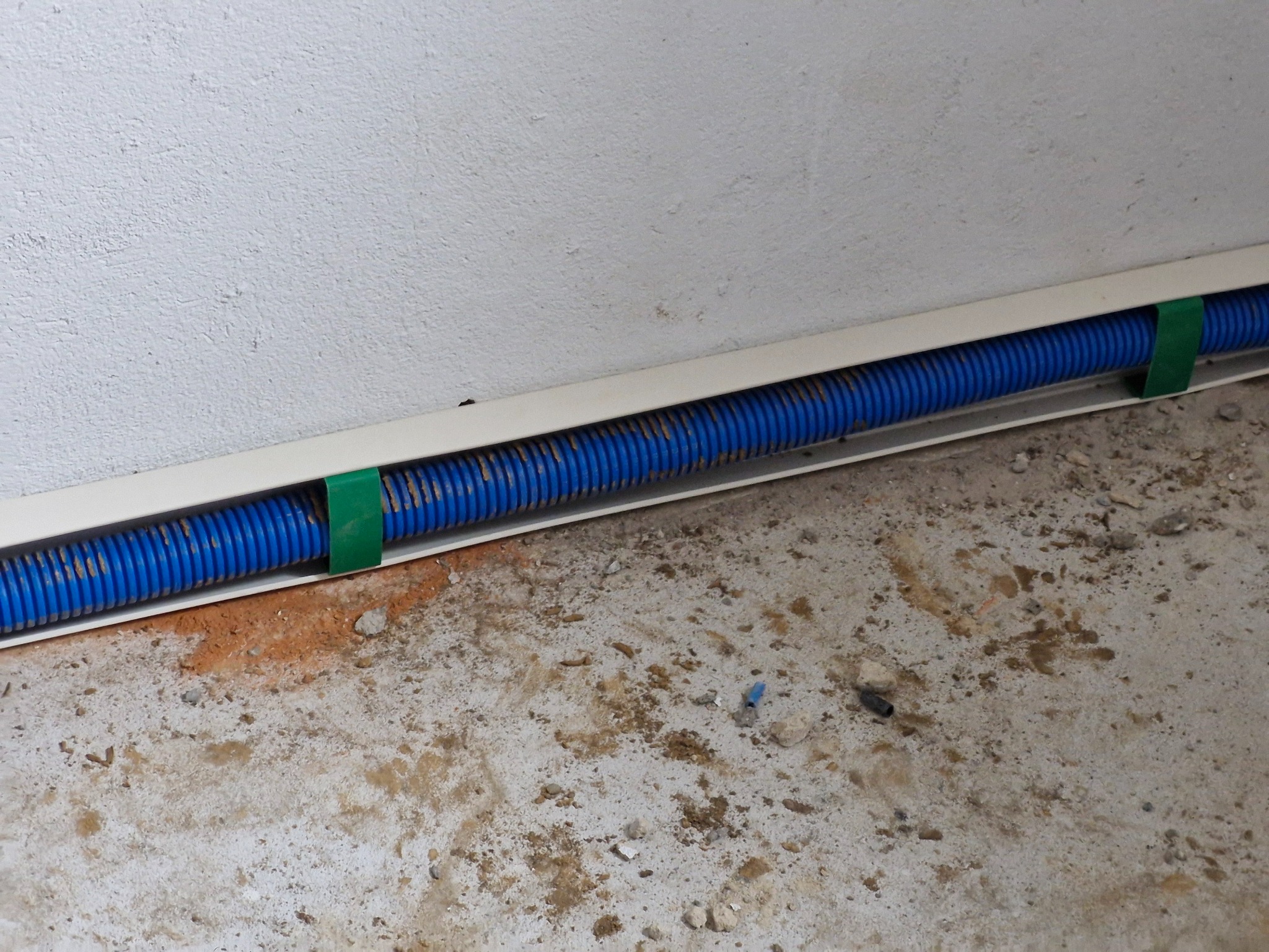 Niebieska rura karbowana w białej listwie przypodłogowej, przymocowana zielonymi uchwytami do ściany. Widoczny surowy beton na podłodze.