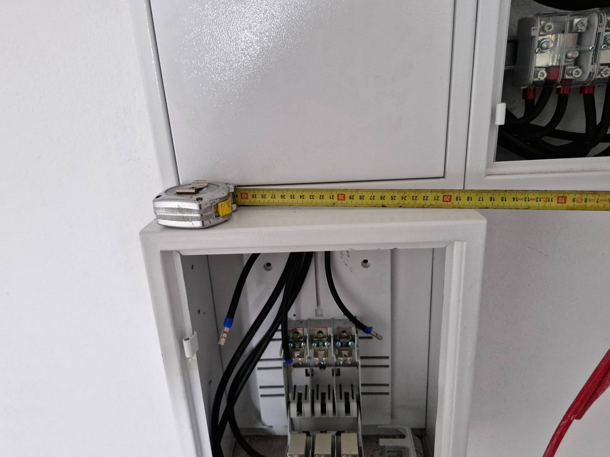Otwarta skrzynka elektryczna z przewodami i listwą zaciskową, pomiar odległości taśmą mierniczą. Widoczne zabezpieczenia nad skrzynką. Białe tło.