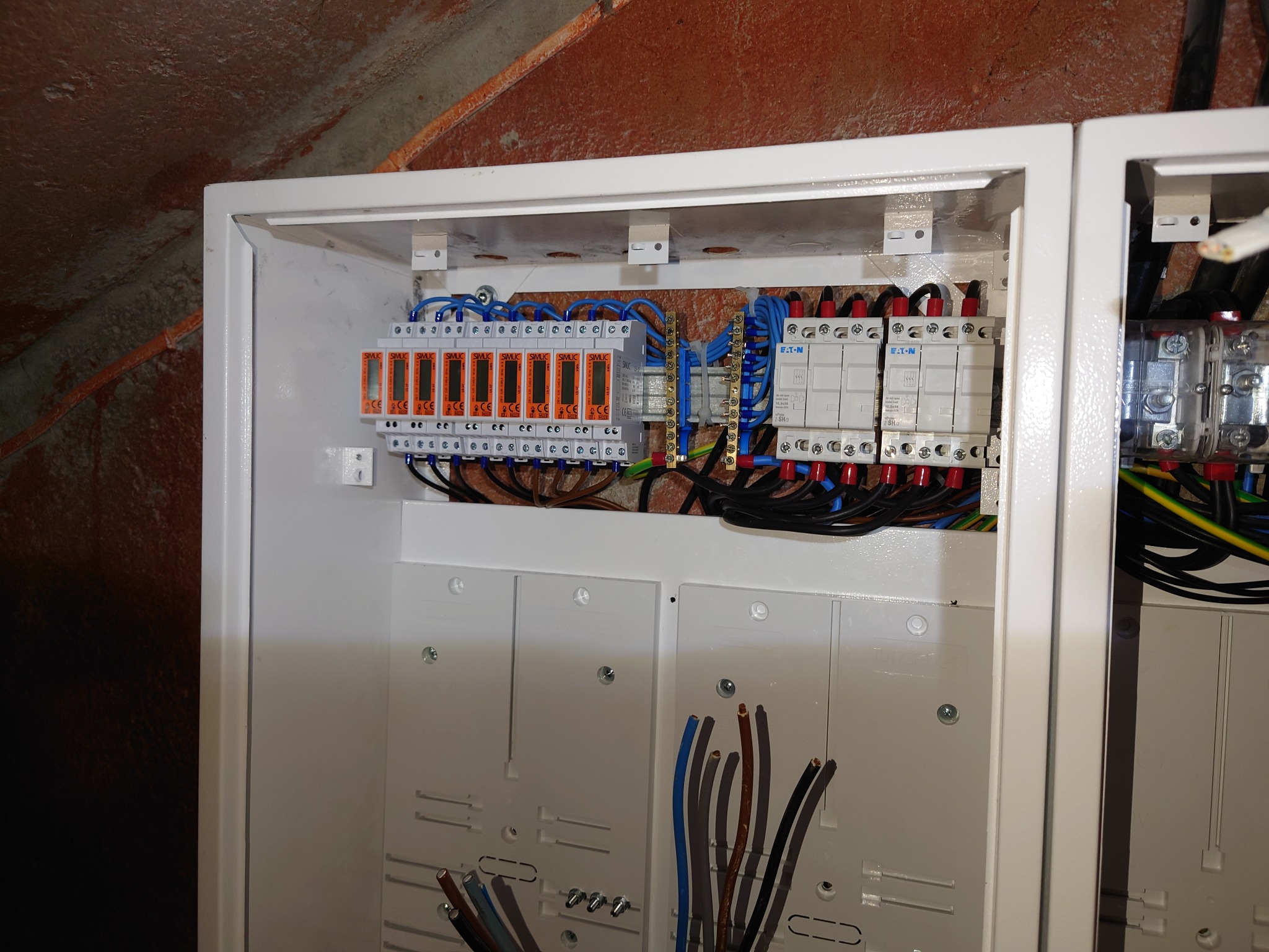 Rozdzielnia elektryczna w białej obudowie, widoczne zabezpieczenia przepięciowe SIMLIC i wyłączniki Eaton, kable w różnych kolorach. Instalacja w trakcie montażu.