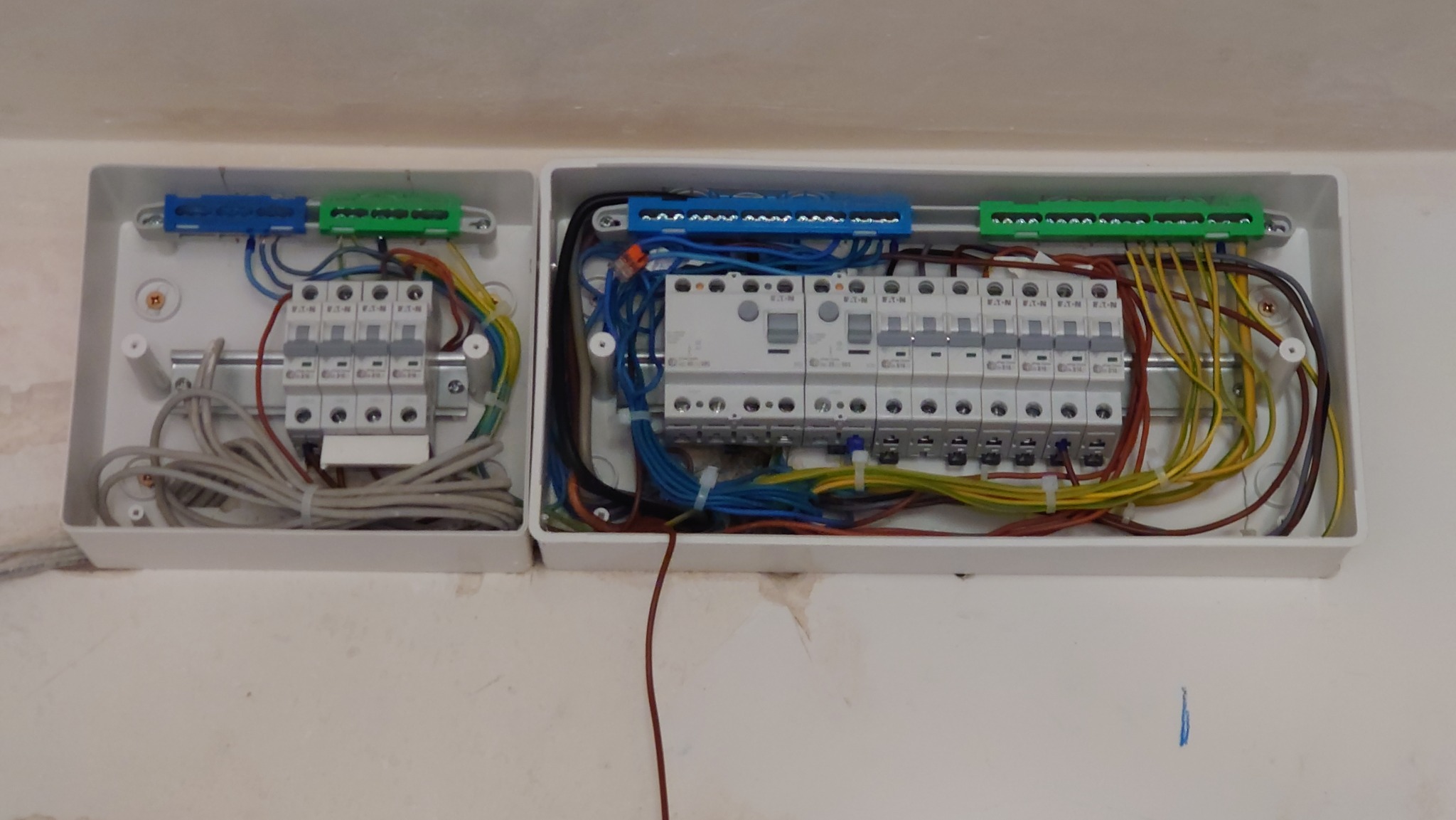 Dwie białe skrzynki elektryczne z zainstalowanymi bezpiecznikami i okablowaniem w kolorach: niebieskim, zielonym, żółtym i brązowym. Widoczne ślady po malowaniu na ścianie.