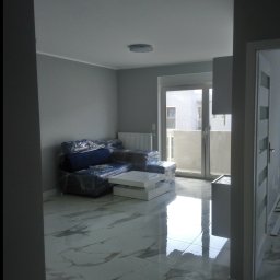 Robert Bińko usługi remontowo wykonczeniowe - Jasne wnętrze po remoncie: marmurowe płytki podłogowe z połyskiem, ściany w odcieniach szarości, meble zabezpieczone folią. Widok na balkon przez duże okno.