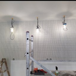 Robert Bińko usługi remontowo wykonczeniowe - Układanie płytek w pionowy wzór w mieszkaniu we Wrocławiu. Widoczne rusztowanie i narzędzia, tymczasowe oświetlenie. Praca glazurnicza w toku.