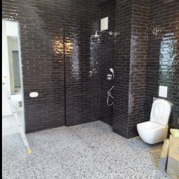 Robert Bińko usługi remontowo wykonczeniowe - Nowoczesna łazienka z czarnymi, błyszczącymi płytkami imitującymi cegłę, szarym podłożem z drobnymi kamyczkami i białym WC. Widoczny prysznic i drabina.