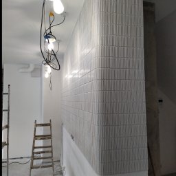 Robert Bińko usługi remontowo wykonczeniowe - Ściana wykończona pionowymi, wypukłymi płytkami w odcieniach szarości, z zaokrąglonym narożnikiem. Widoczne kable i lampy wiszące, drabina. Wnętrze w trakcie prac.