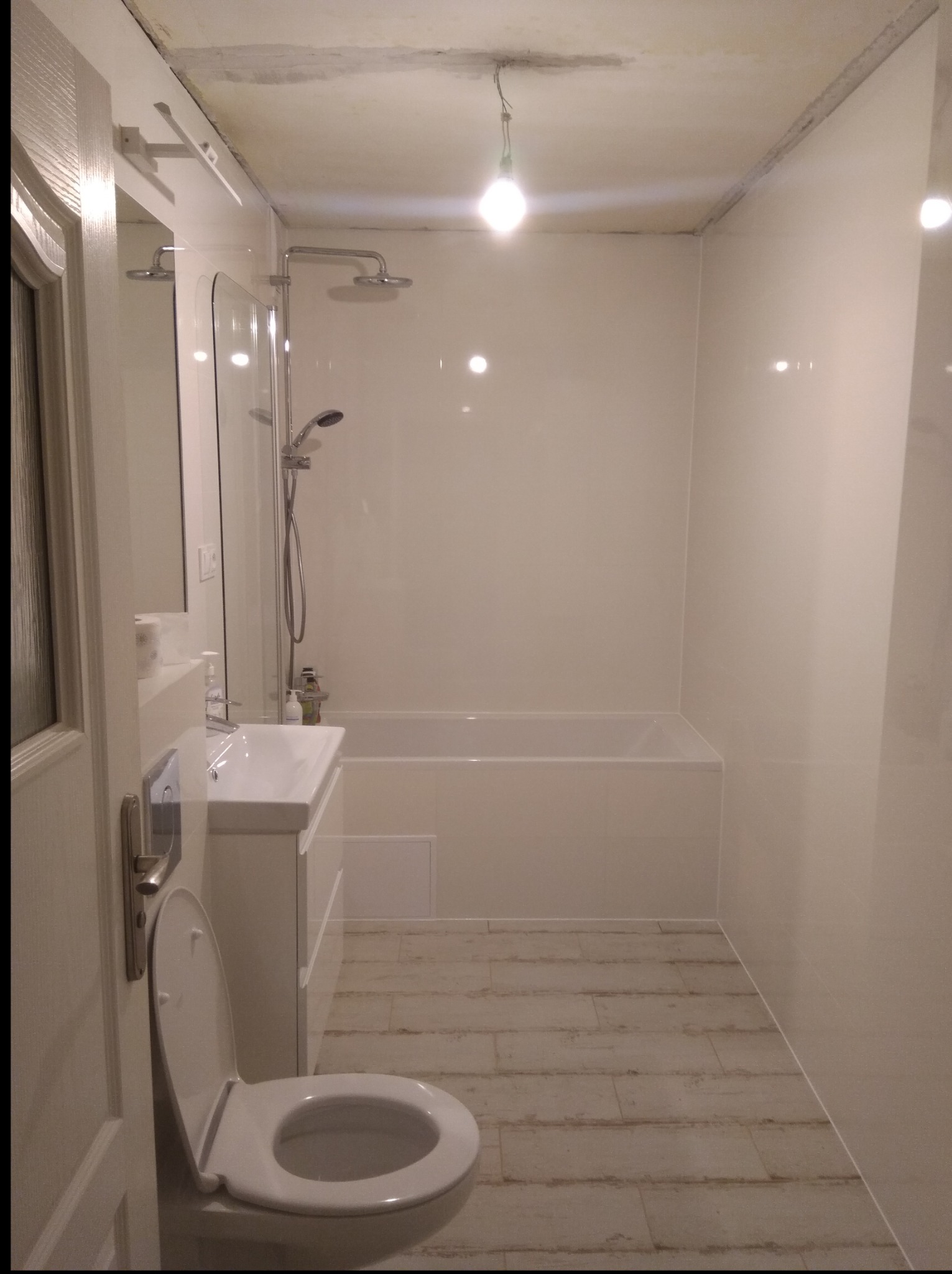 Biała łazienka po remoncie z wanną, umywalką i toaletą. Widoczne płytki podłogowe imitujące drewno. Sufit w trakcie wykończenia. Minimalistyczny styl.