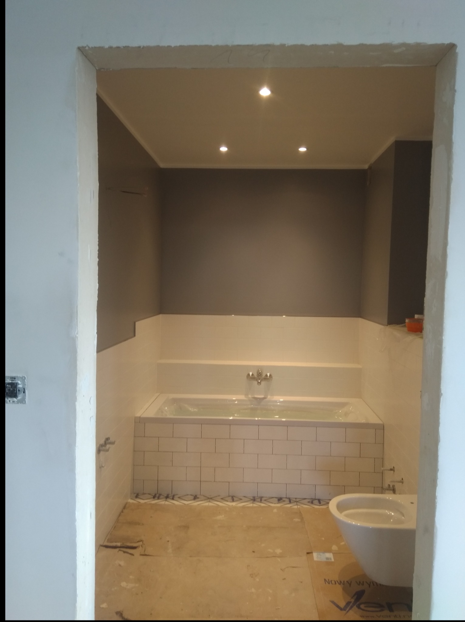 Łazienka w trakcie remontu: białe płytki na wannie i ścianach, szara ściana, częściowo zamontowana toaleta, widoczne niedoskonałości wykończenia ścian i podłogi.