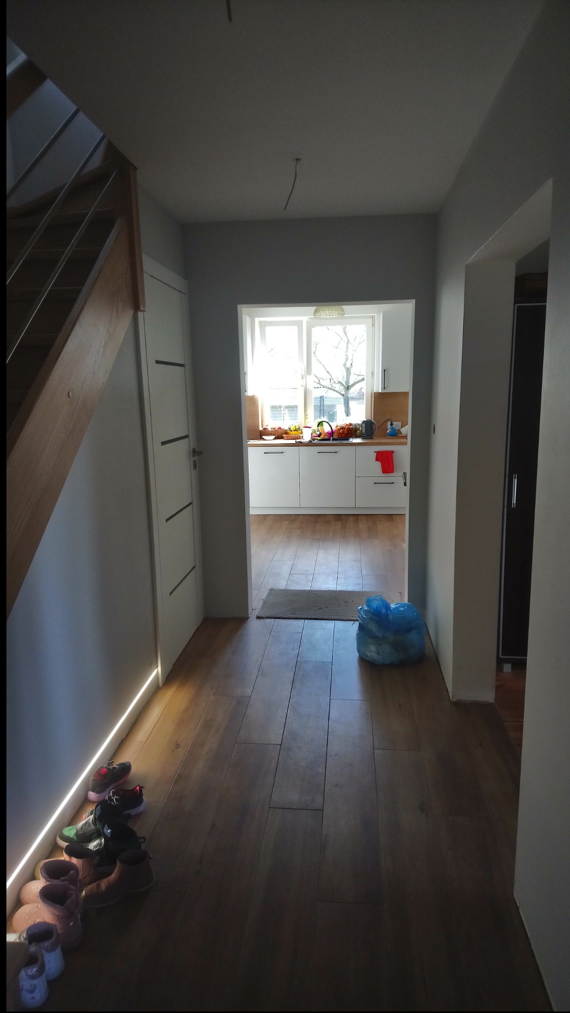 Jasny korytarz po remoncie z drewnianą podłogą, białymi ścianami i widokiem na kuchnię w tle. W rogu schody i buty. Widać ślady po szpachlowaniu.