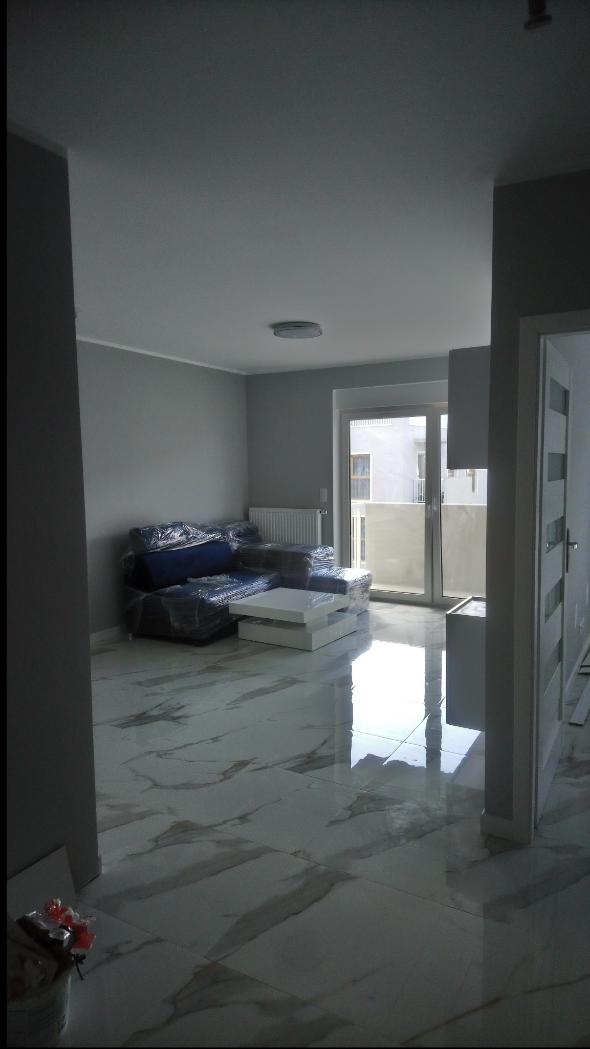 Jasne wnętrze po remoncie: marmurowe płytki podłogowe z połyskiem, ściany w odcieniach szarości, meble zabezpieczone folią. Widok na balkon przez duże okno.