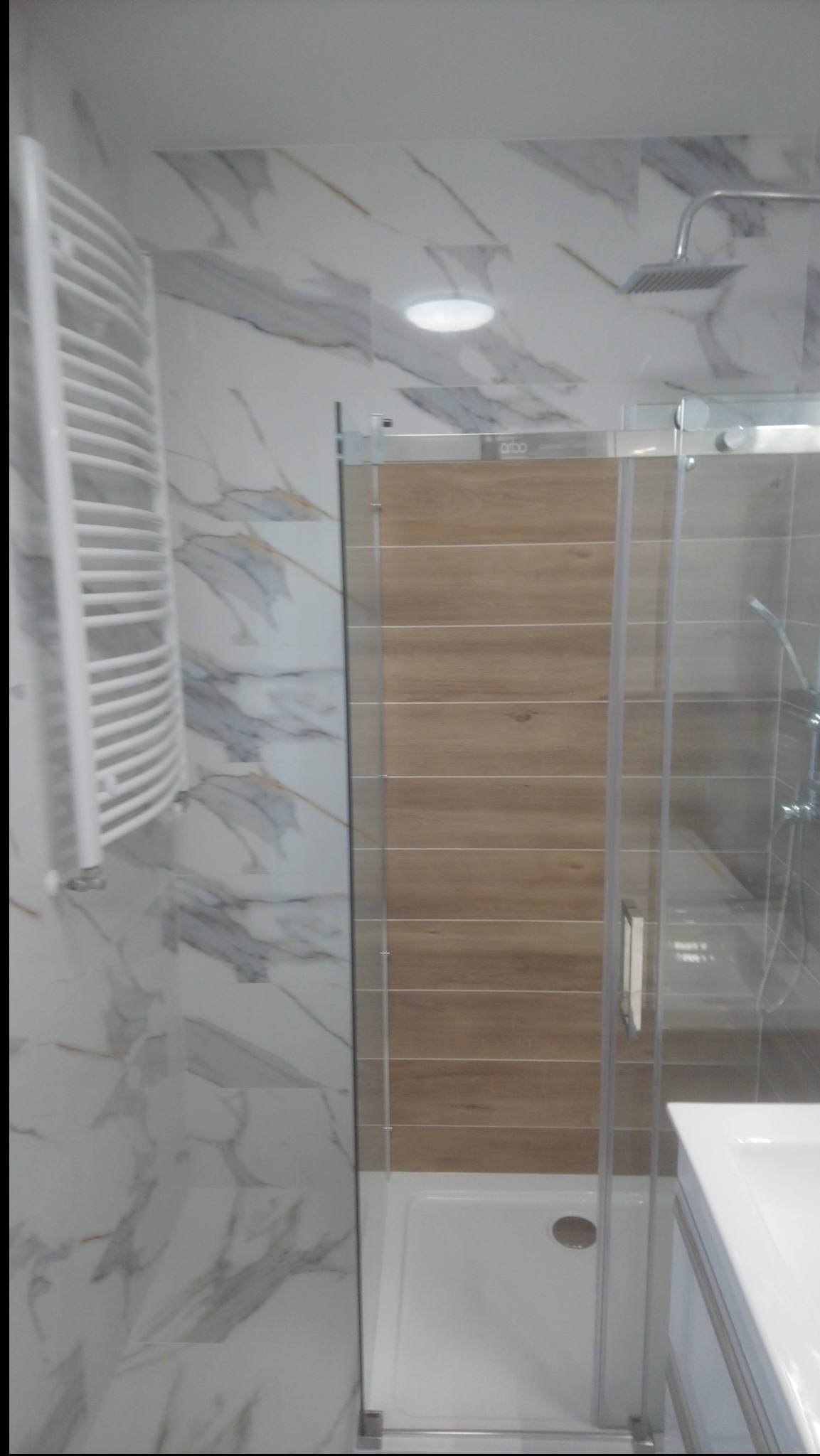 Wykończona łazienka z kabiną prysznicową z drewnianym panelem, grzejnikiem drabinkowym i marmurowymi płytkami. Nowoczesny design, jasne oświetlenie.