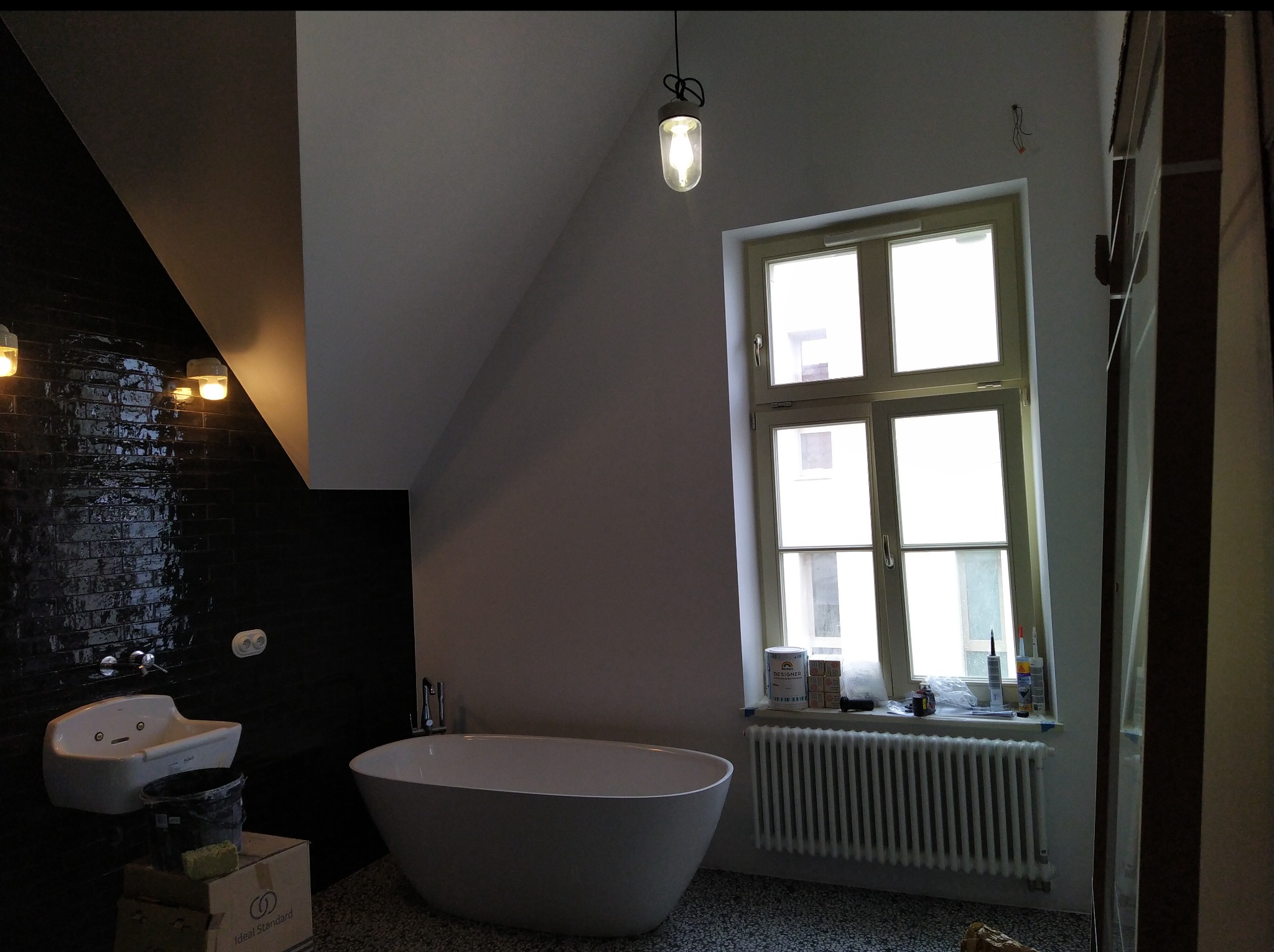 Biała wanna wolnostojąca i umywalka w łazience z czarnymi płytkami, okno z grzejnikiem. W tle skośny sufit i wisząca lampa. Widoczne akcesoria i materiały budowlane.