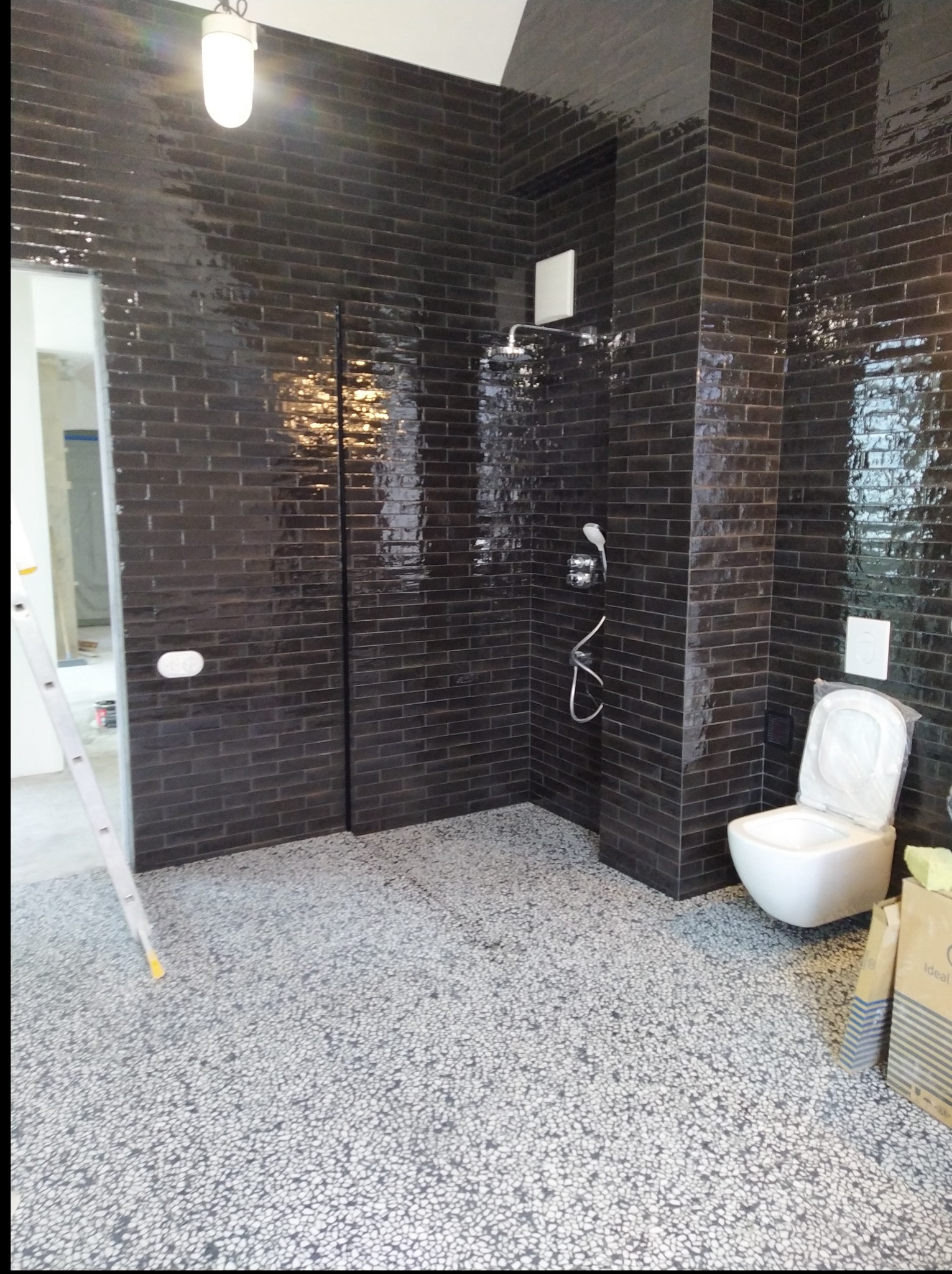 Nowoczesna łazienka z czarnymi, błyszczącymi płytkami imitującymi cegłę, szarym podłożem z drobnymi kamyczkami i białym WC. Widoczny prysznic i drabina.