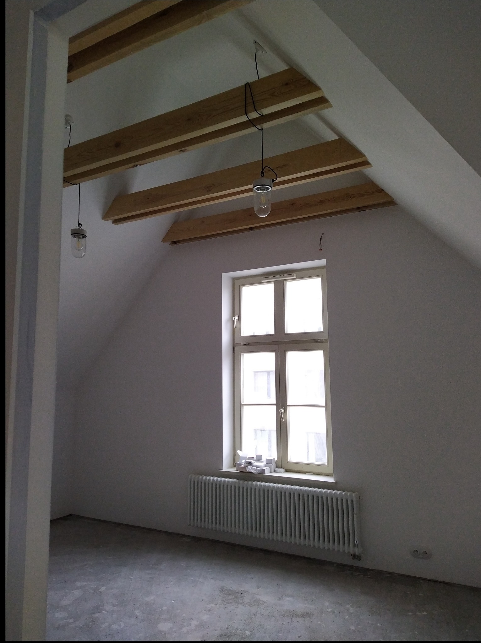Poddasze w trakcie remontu: surowe drewniane belki stropowe, okno z grzejnikiem, surowy beton na podłodze, wiszące żarówki. Ściany pomalowane na biało.