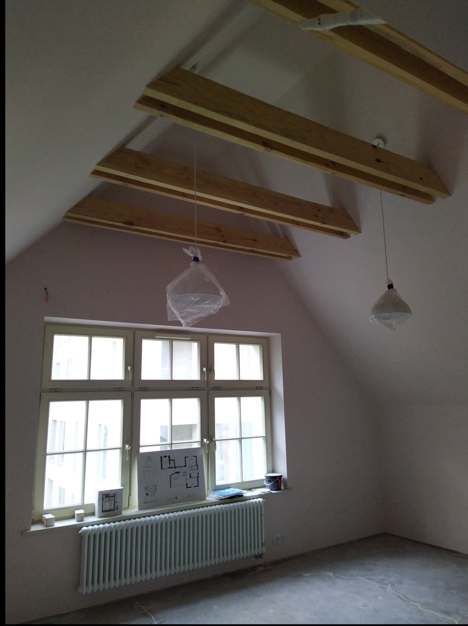 Poddasze w trakcie prac wykończeniowych: surowe drewniane belki pod stropem, okno z parapetem, grzejnik, plan mieszkania i wiszące lampy w folii ochronnej.