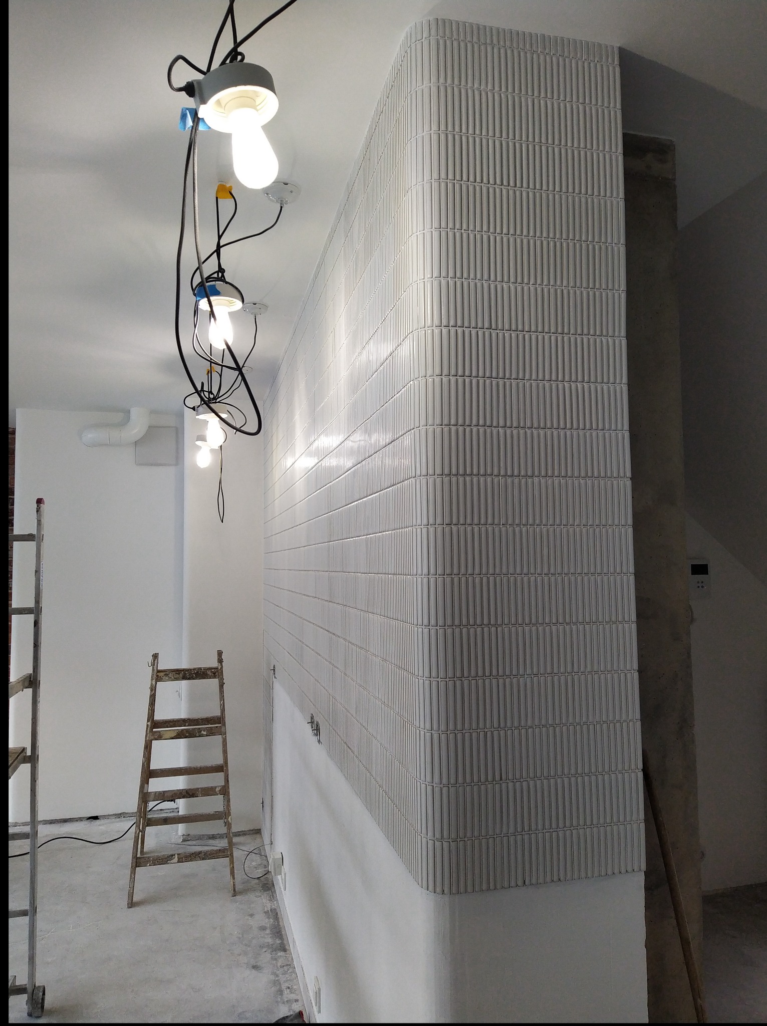 Ściana wykończona pionowymi, wypukłymi płytkami w odcieniach szarości, z zaokrąglonym narożnikiem. Widoczne kable i lampy wiszące, drabina. Wnętrze w trakcie prac.