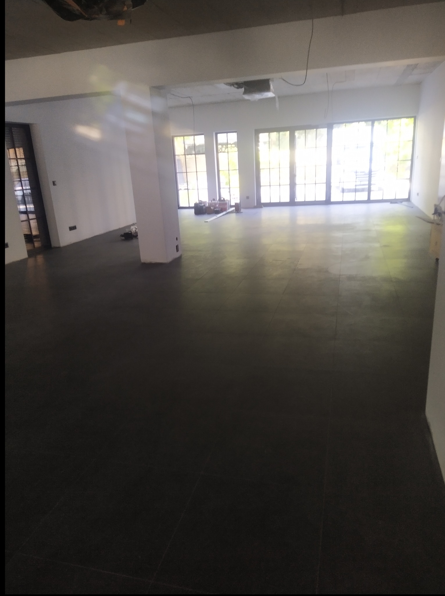 Ukończona, ciemna posadzka w przestronnym wnętrzu z dużymi oknami. Widoczne narzędzia i materiały budowlane w tle, sugerujące trwające prace wykończeniowe.