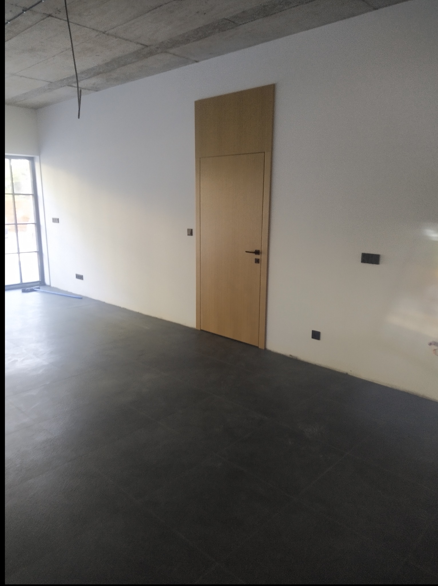 Wnętrze w trakcie wykańczania: ciemna podłoga, białe ściany z wystającymi kablami i zamontowane drzwi z ościeżnicą w kolorze jasnego drewna.