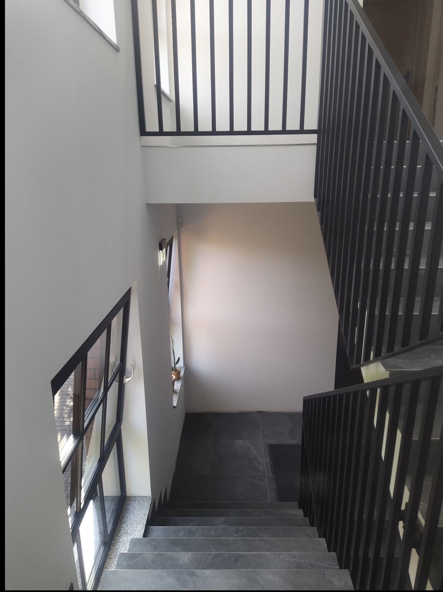 Klatka schodowa z szarymi schodami i czarną balustradą. Widok z góry na dół, okno po lewej stronie. Minimalistyczne wykończenie wnętrza, ciemna podłoga na podeście.
