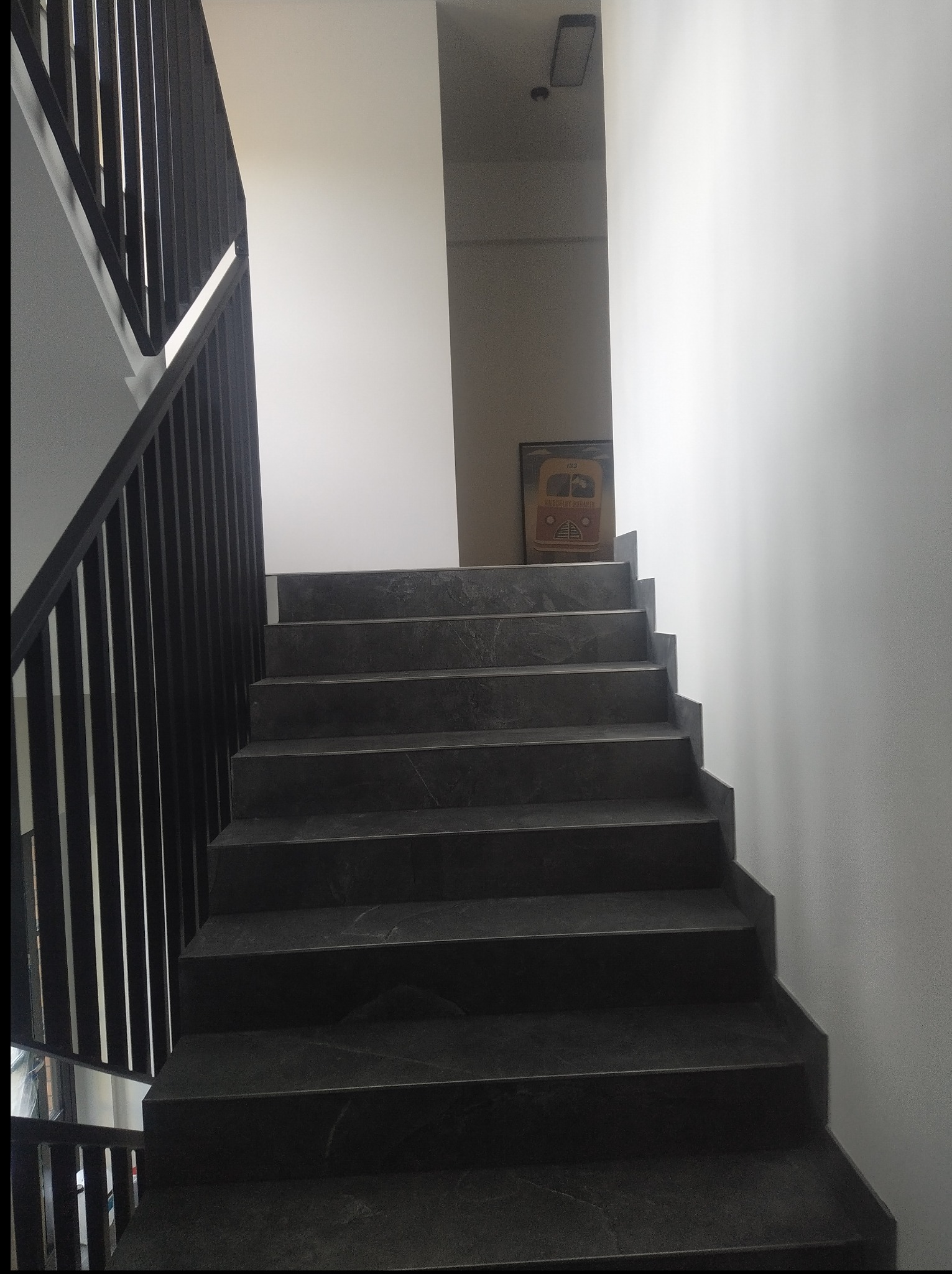 Ciemne, marmurowe schody z czarną, metalową balustradą w nowoczesnym wnętrzu. Na ścianie widoczny obrazek autobusu, oświetlony górnym światłem.