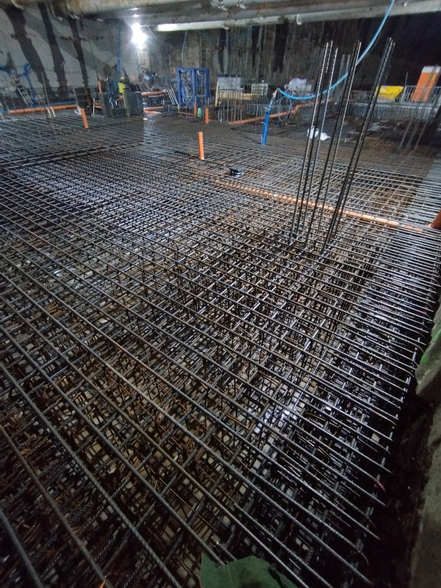 Zbrojenie fundamentu z wystającymi prętami, widok z góry na siatkę zbrojeniową, rury instalacyjne w tle, surowy stan budowy.