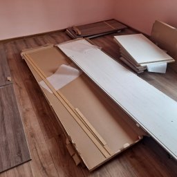 Usługi budowlane elektryczne - Rozpakowane elementy mebli z płyty wiórowej na podłodze z paneli. Widoczne kartony, listwy zabezpieczające i akcesoria montażowe. Przygotowanie do montażu.