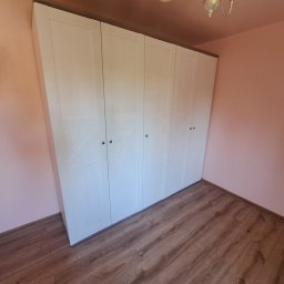 Usługi budowlane elektryczne - Biała, czterodrzwiowa szafa z ciemnobrązowym blatem, zamontowana w pokoju z różowymi ścianami i drewnianą podłogą. Widoczne detale wykończenia drzwi.