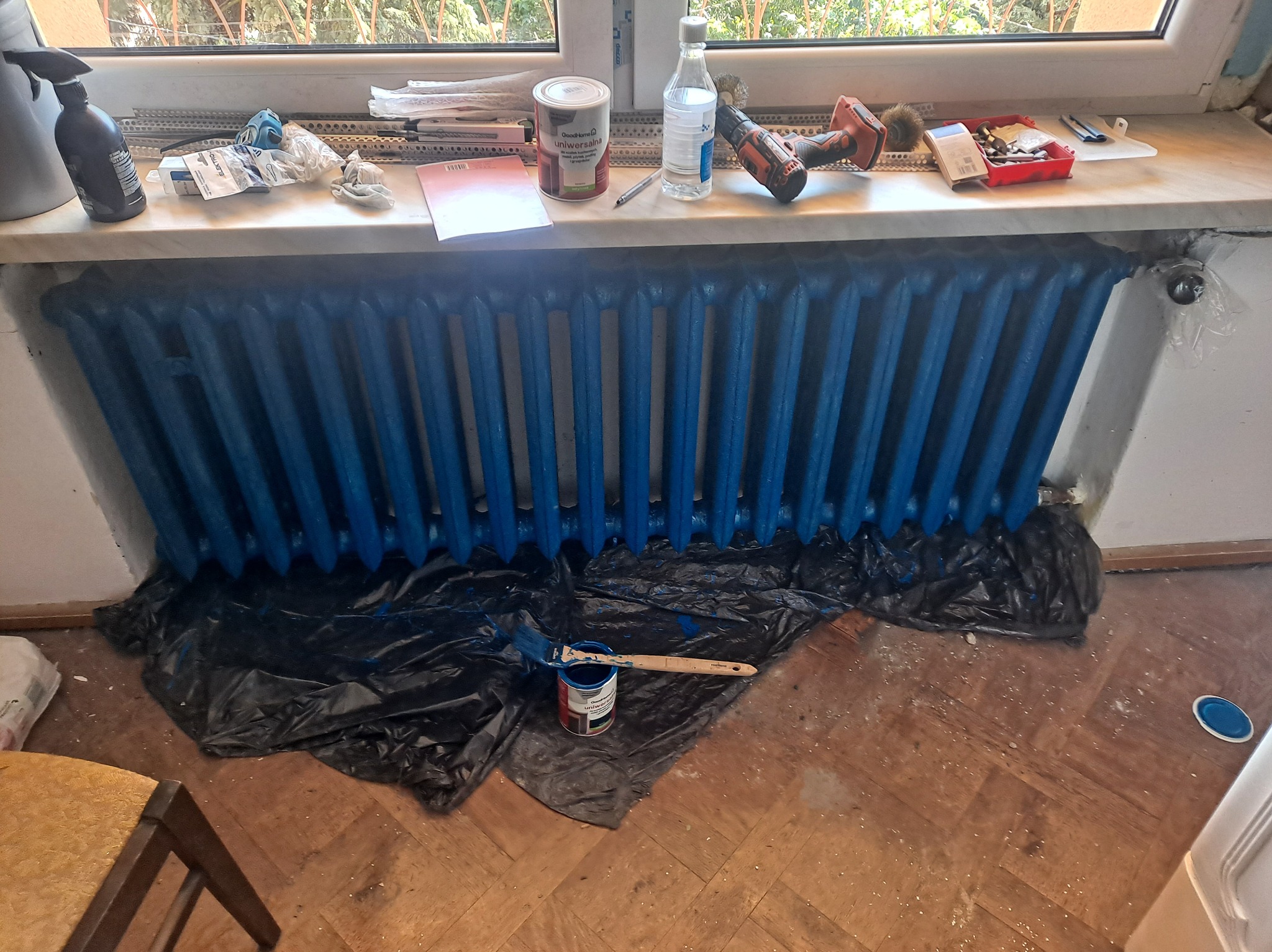 Malowanie żeliwnego grzejnika na niebiesko. Pędzel i farba na folii ochronnej. Na parapecie narzędzia i akcesoria malarskie. Widok z góry.