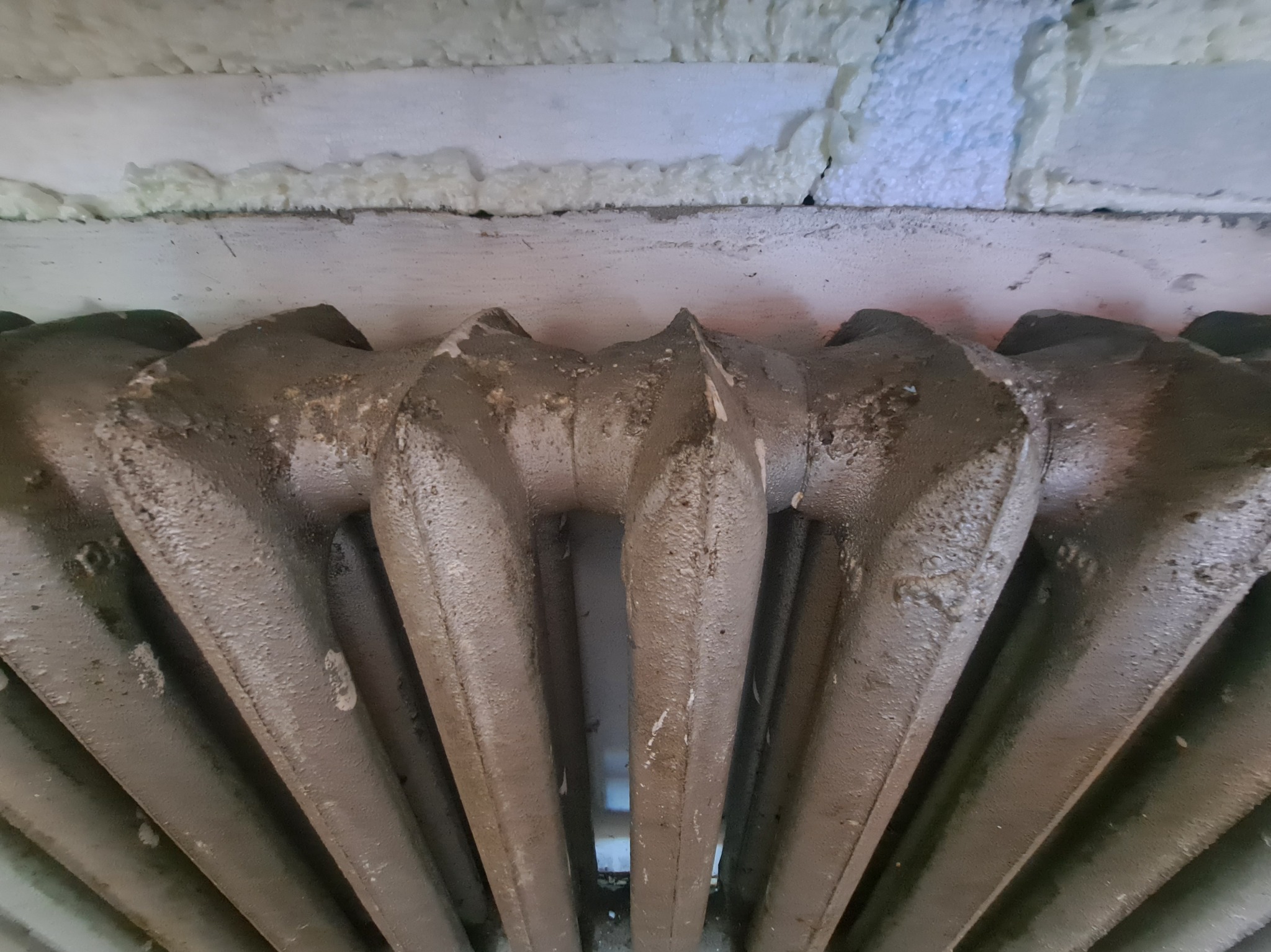 Stary, żeliwny grzejnik pomalowany na szaro, z widoczną pianką izolacyjną w tle. Ukazany z bliska, z perspektywy od dołu, z detalami korozji i nierówności farby.