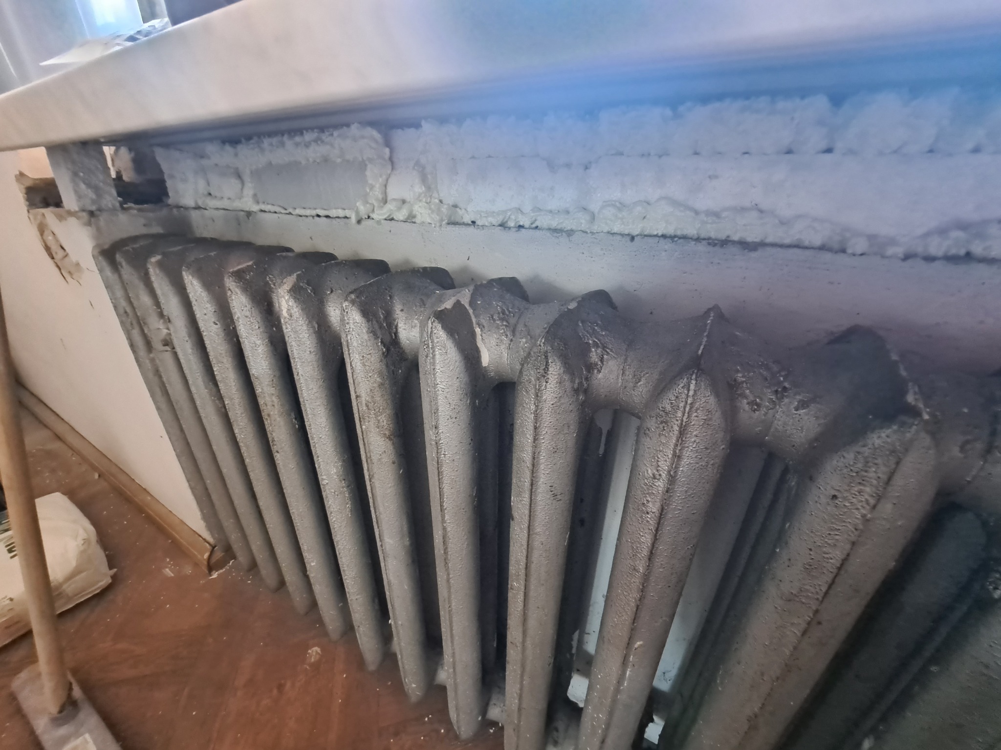 Szczeliny pod parapetem wypełnione pianką montażową, poniżej stary, żeliwny grzejnik. Prace remontowe w trakcie uszczelniania instalacji grzewczej.