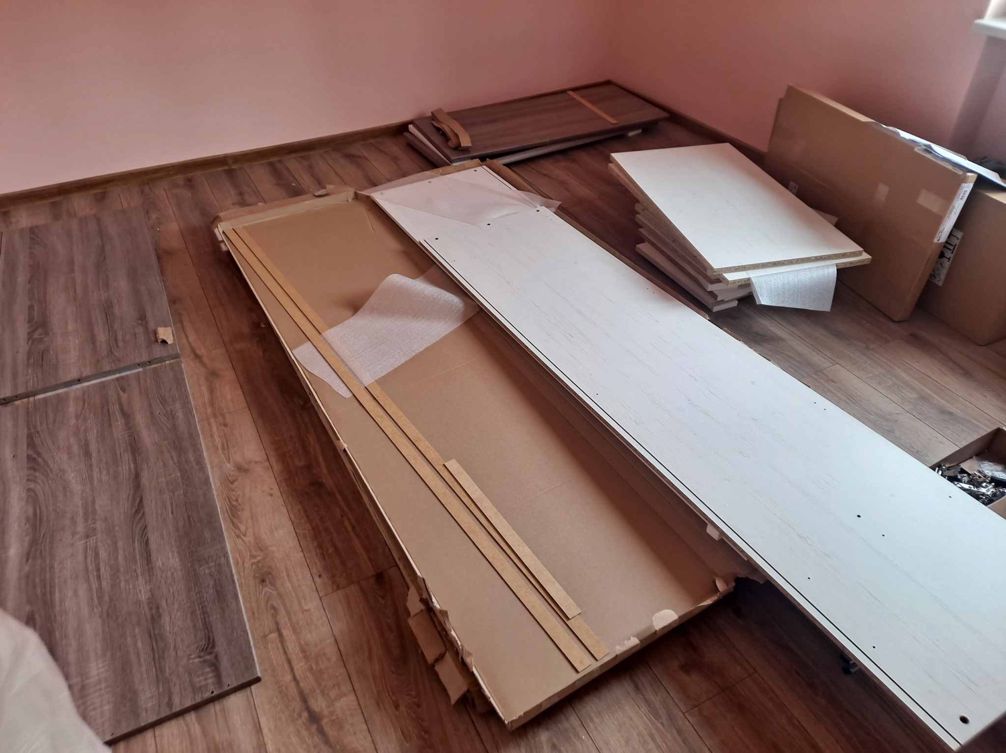 Rozpakowane elementy mebli z płyty wiórowej na podłodze z paneli. Widoczne kartony, listwy zabezpieczające i akcesoria montażowe. Przygotowanie do montażu.
