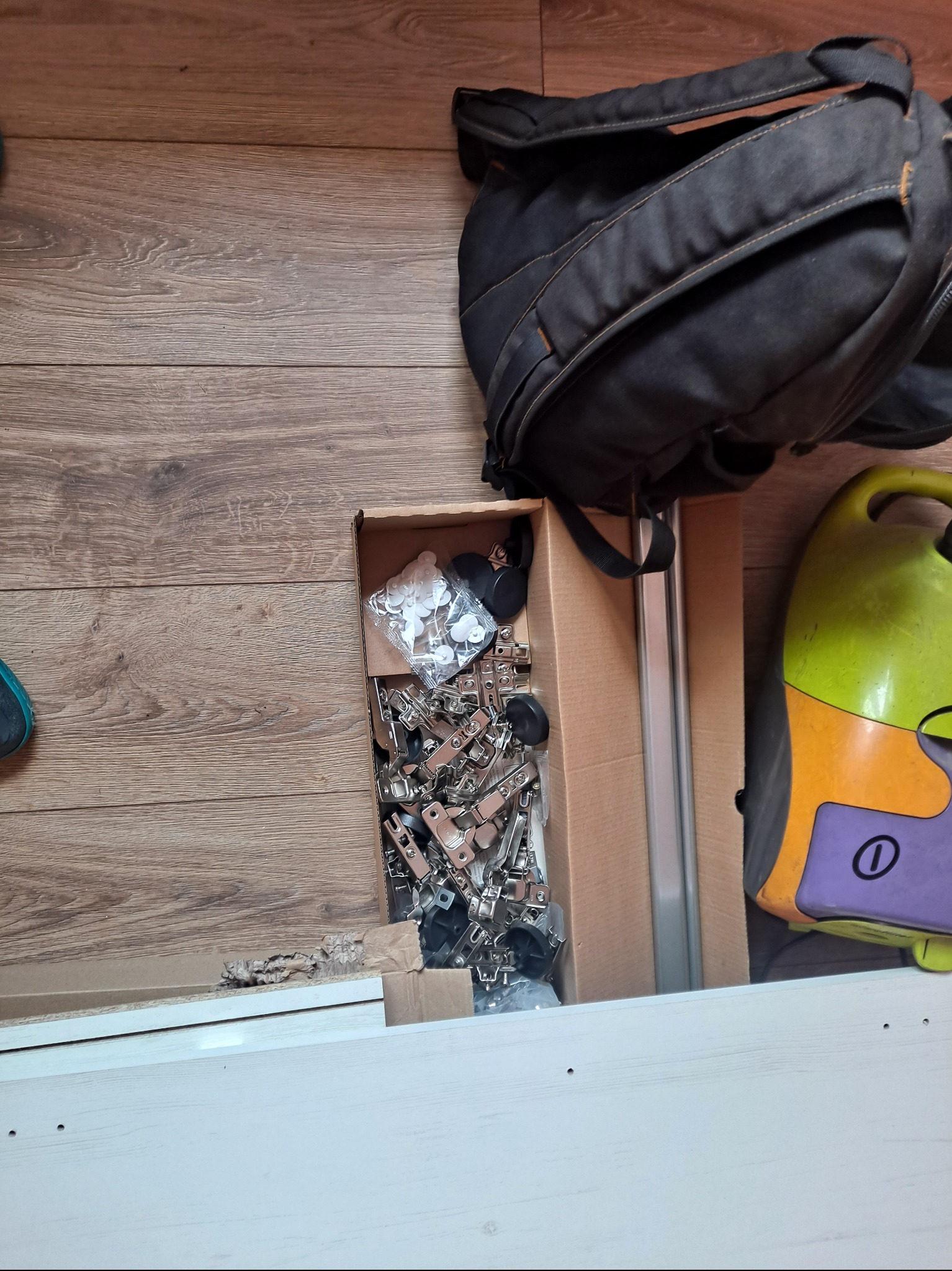 Rozpakowane okucia meblowe w kartonie na podłodze z panelem, obok plecak i odkurzacz. Przygotowanie do montażu mebli z białej płyty.