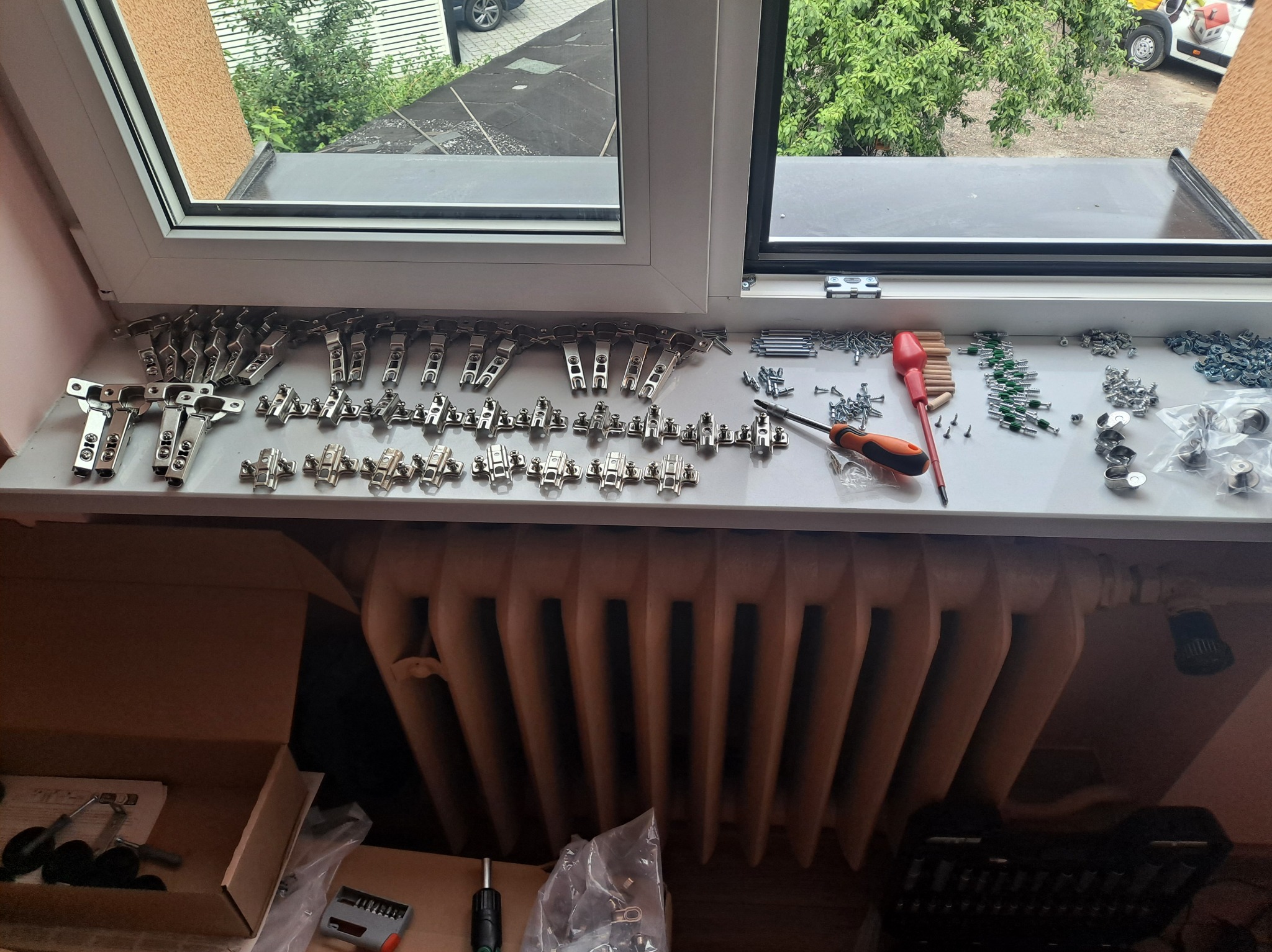 Przygotowane zawiasy, śruby i narzędzia na parapecie okna, gotowe do montażu drzwi szafki. Widoczny grzejnik i kartony z elementami. Ujęcie z góry.