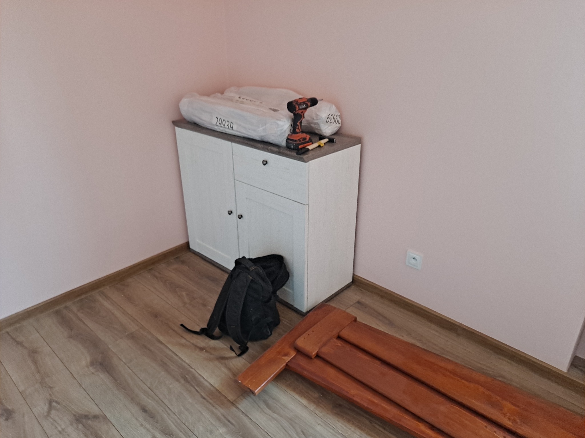 Biała szafka z szarą górą, wiertarka, zapakowane elementy mebli, plecak i fragmenty drewnianej konstrukcji na podłodze w pokoju z różowymi ścianami.