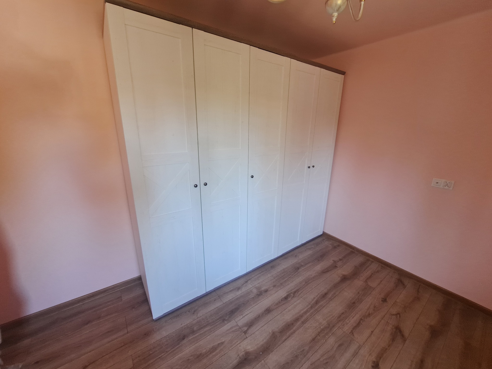 Biała, czterodrzwiowa szafa z ciemnobrązowym blatem, zamontowana w pokoju z różowymi ścianami i drewnianą podłogą. Widoczne detale wykończenia drzwi.