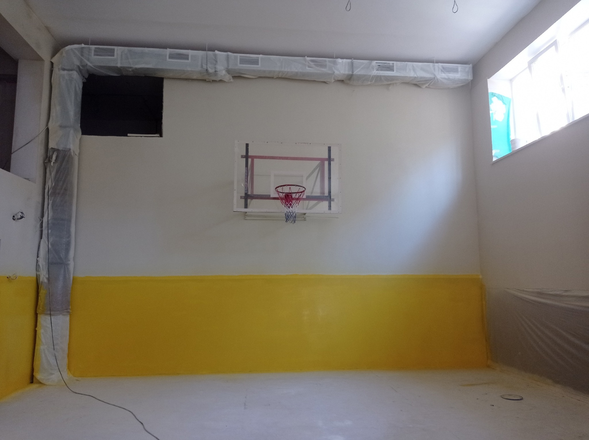 Świeżo pomalowana sala gimnastyczna z żółtą lamperią i białymi ścianami. Elementy instalacji zabezpieczone folią malarską. Widoczny kosz do koszykówki.
