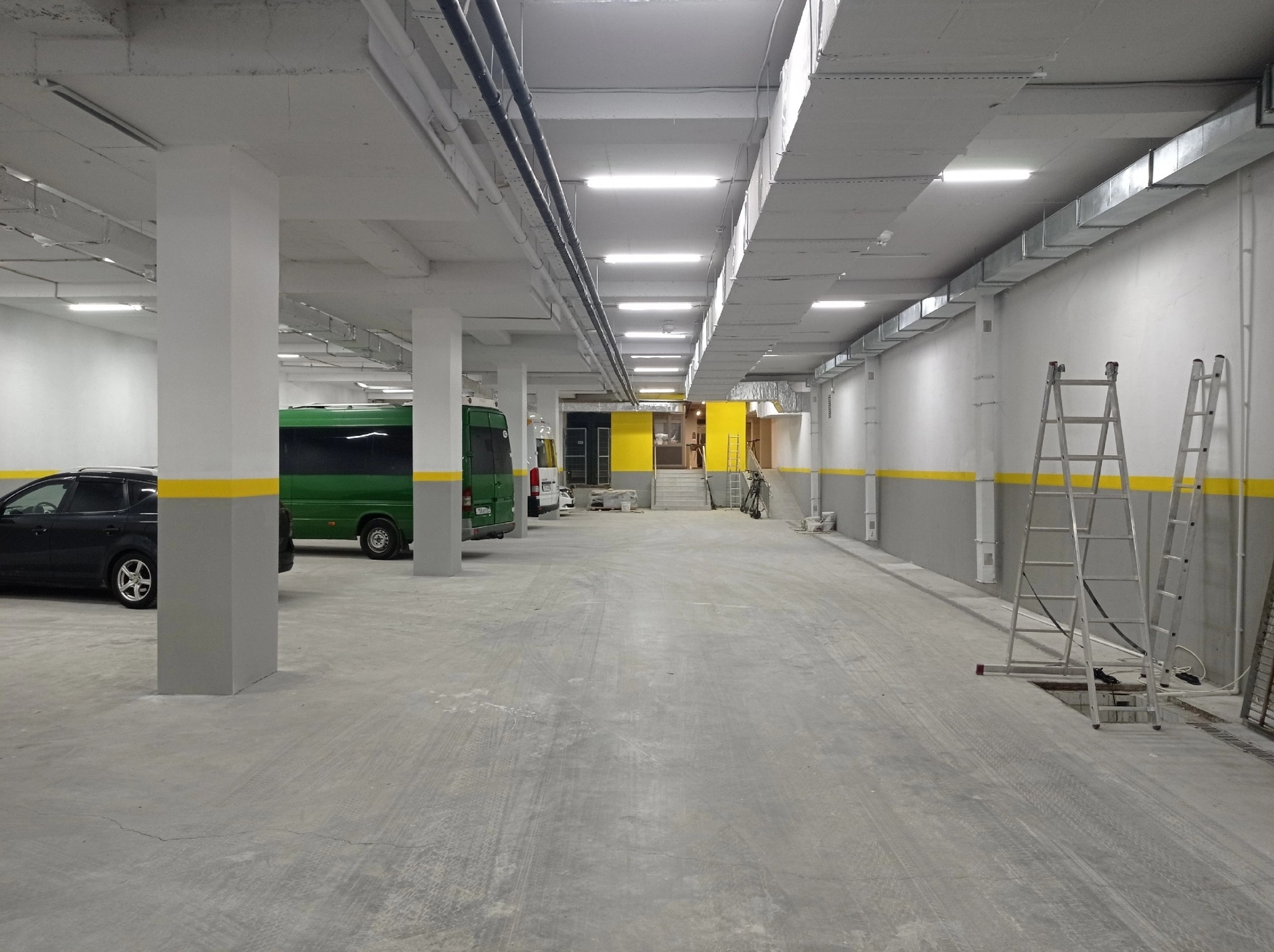 Wnętrze parkingu podziemnego po remoncie: świeżo pomalowane ściany w szaro-białej tonacji z żółtym pasem, widoczne słupy, instalacje pod sufitem i drabiny.