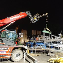 SPAW-BUD - Czerwony dźwig Manitou podnosi zieloną płytę na budowie nocą, widoczne rusztowania i pracownik w kabinie.