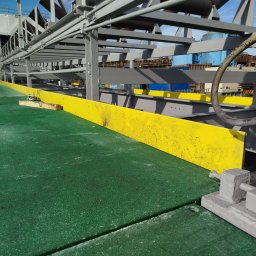 SPAW-BUD - Konstrukcja stalowa z żółtą osłoną na zielonej platformie, widoczny pracownik w oddali. Metalowa instalacja z kablami i elementami mocującymi.