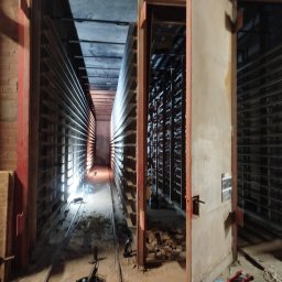 SPAW-BUD - Wnętrze komory suszarniczej z metalowymi regałami, widoczne narzędzia i resztki materiałów budowlanych, oświetlenie punktowe w głębi. Drzwi komory otwarte.