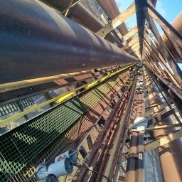 SPAW-BUD - Widok z dołu na metalową konstrukcję z rurami i kratownicami, z pracownikami na górze. Perspektywa z dołu podkreśla złożoność i wysokość instalacji.