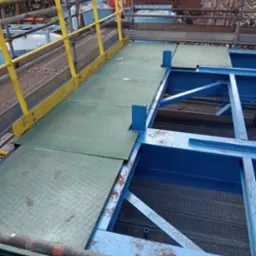 Platforma robocza z żółtą barierką i zielonymi panelami na tle konstrukcji stalowej i rusztowań. Widok z góry na fragment instalacji przemysłowej.