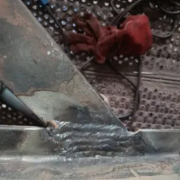 Spoina łącząca metalowe elementy konstrukcyjne, widoczne ślady spawania, obok leży rękawica robocza i narzędzie do obróbki metalu na metalowej perforowanej powierzchni.