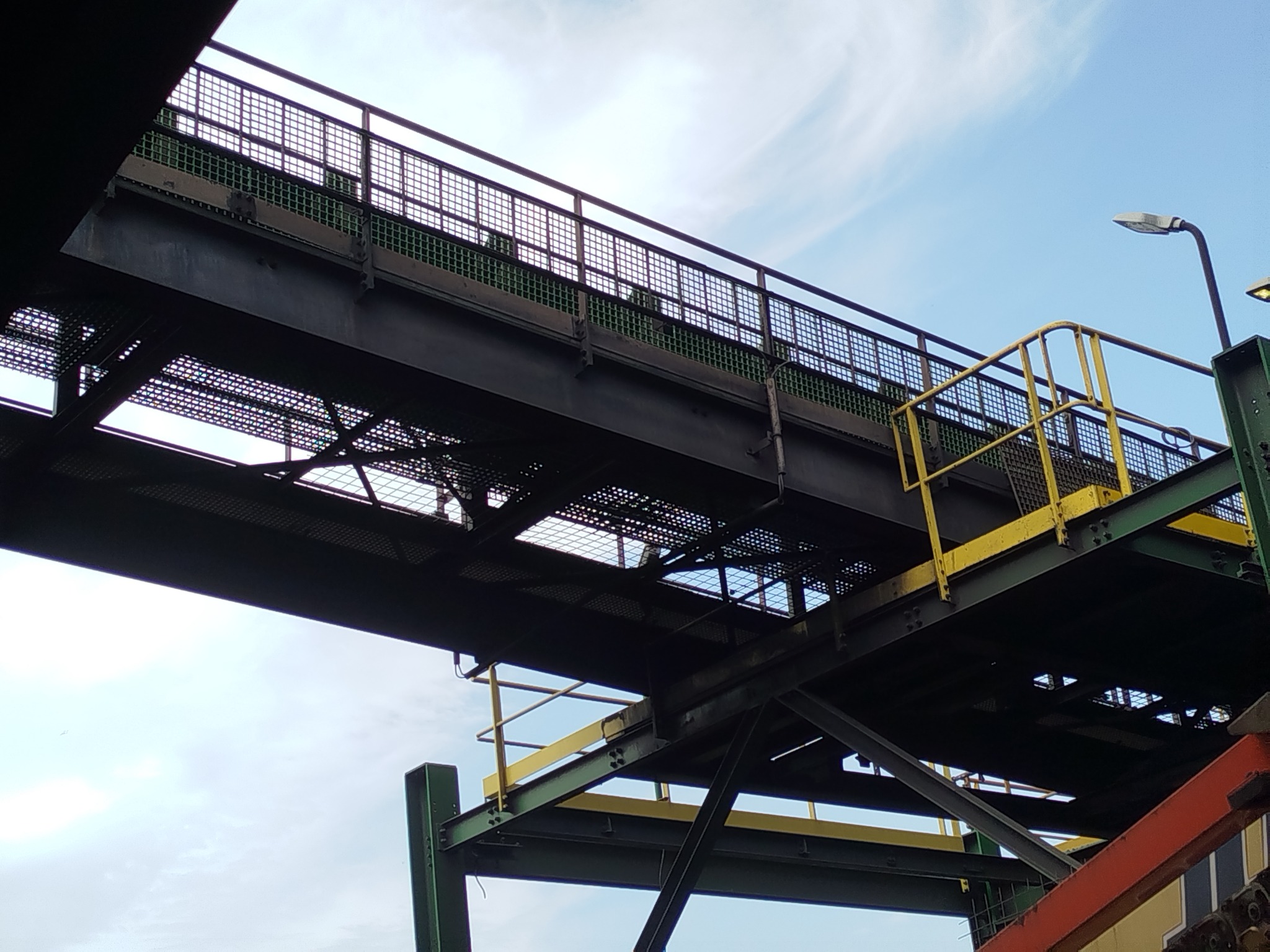 Metalowa konstrukcja pomostu z żółtymi barierkami i oświetleniem ulicznym, widok z dołu na błękitne niebo.