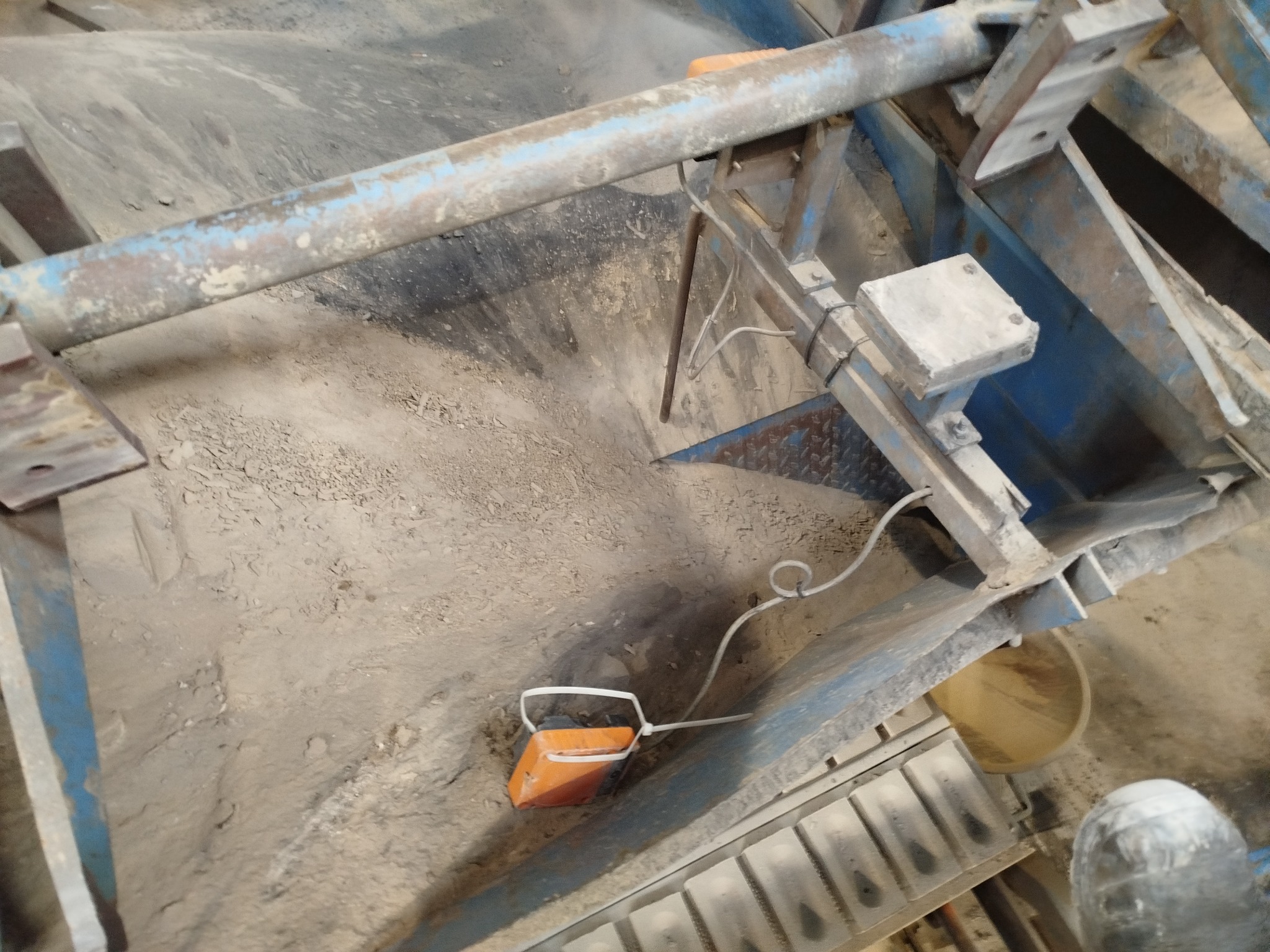 Widok z góry na zanieczyszczoną konstrukcję z metalowymi elementami, przewodami i pomarańczową kostką. Całość w odcieniach szarości i błękitu, z widocznymi śladami użytkowania.